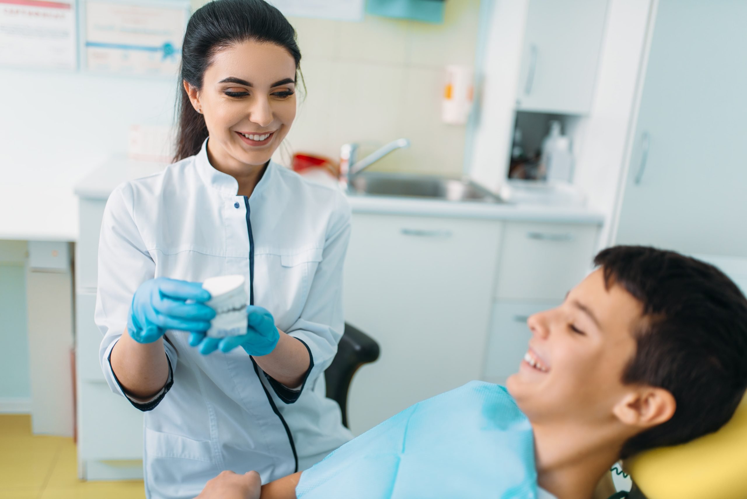 A friendly female dentist is showing a dental model to a young boy. Comprehensive pediatric care from a Falls Church Dentist at Broadway Dental Associates.