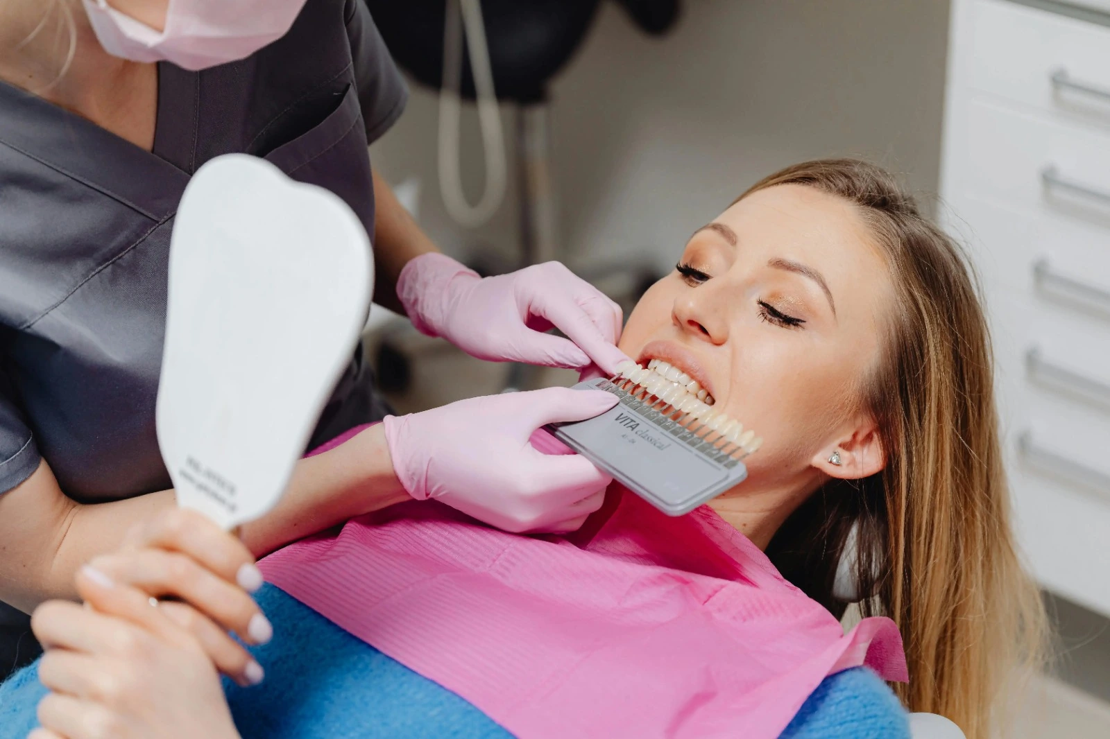 A dentist matches veneers with a patient’s teeth, who is holding a mirror to see them