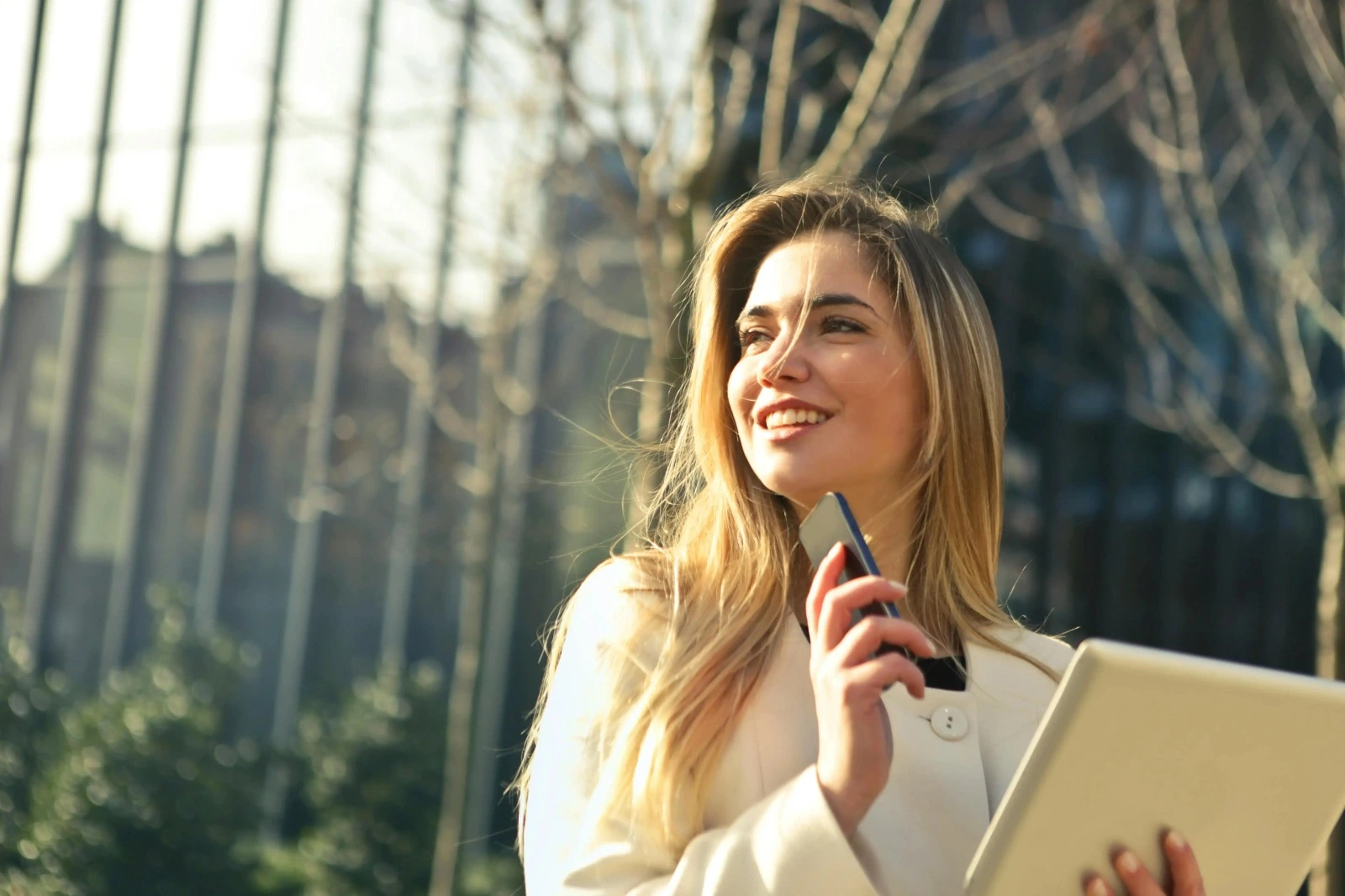 A young woman with blonde hair is smiling while holding her phone with one hand.