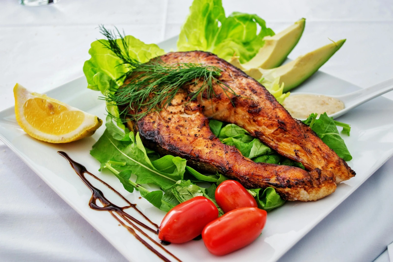 Deliciously fried fish is served on a plate along with it are small tomatoes, lemon, green leaves, and chopped avocados.