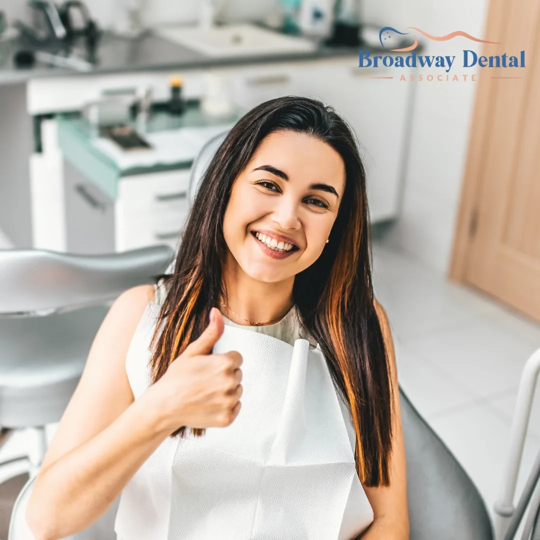 A smiling young woman wearing white, in a dental office, gives a thumbs-up.