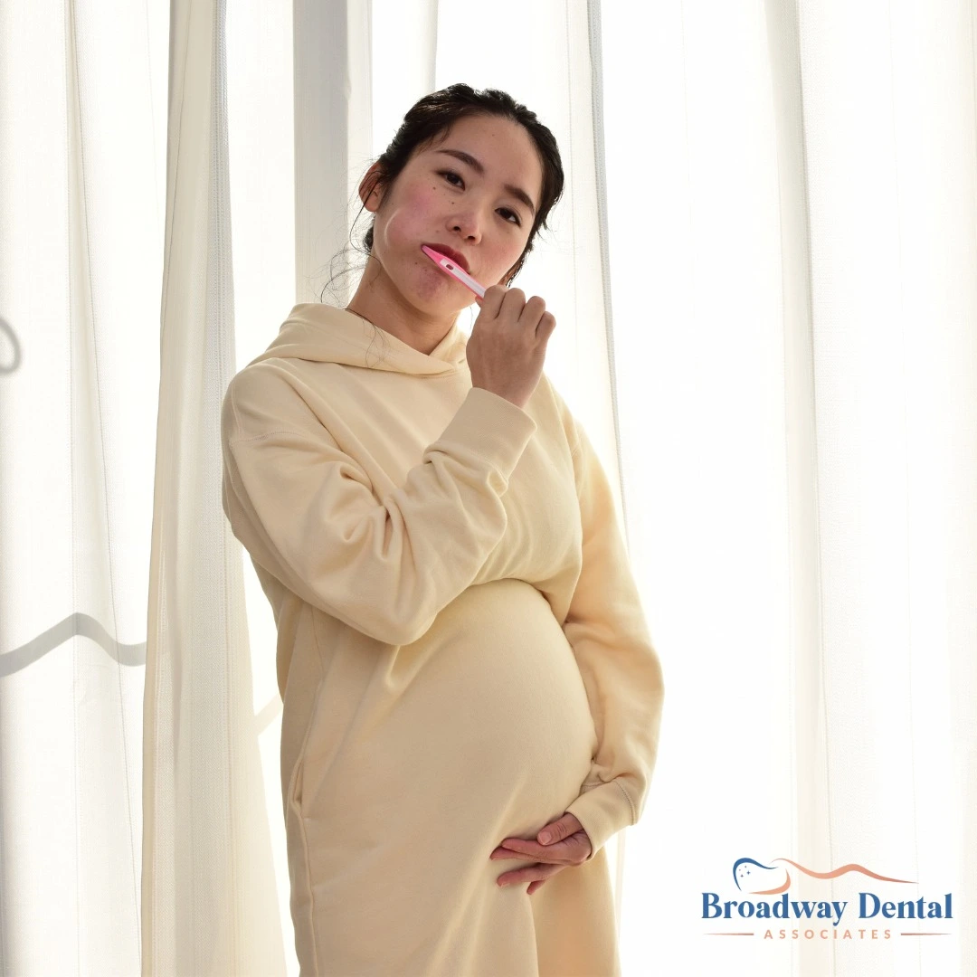 An expectant young woman with dark hair pulled back, wearing a light beige sweatshirt, brushing her teeth in front of a bright window, emphasizing the need to during pregnancy