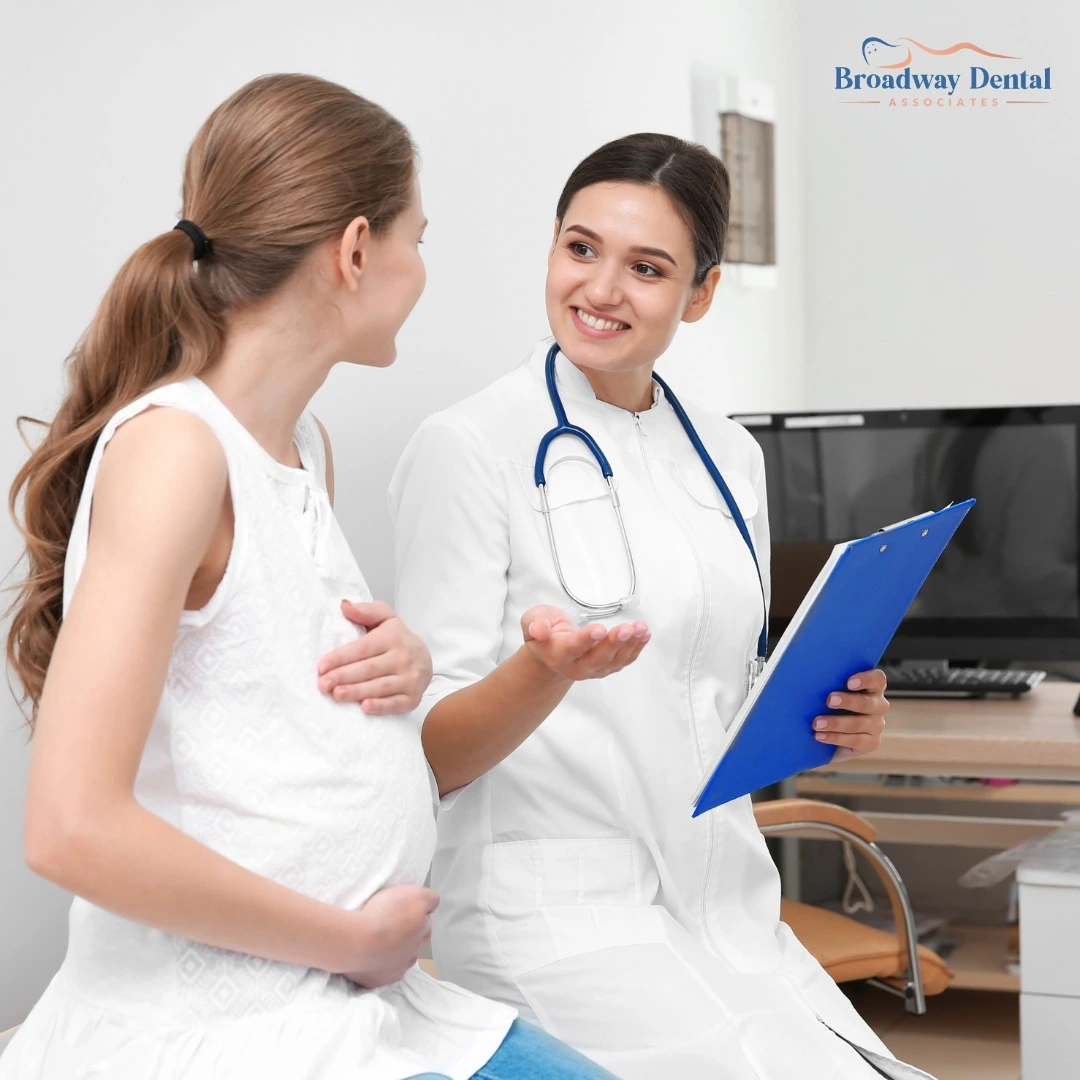 Pregnant woman in a white top smiling at her dentist who is also smiling, wearing a stethoscope and holding a blue clipboard.