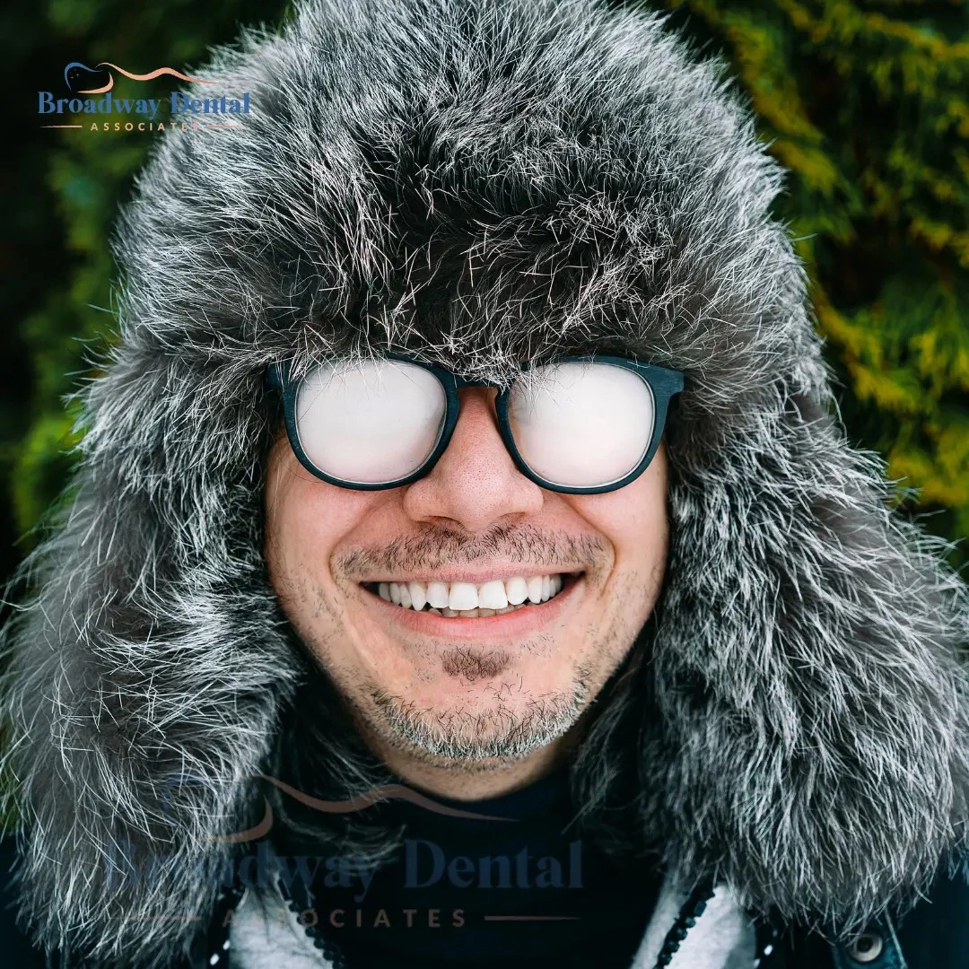Man smiling with bright white teeth in a furry winter hat and fogged-up glasses and Broadway Dental Associates logo at the top left.
