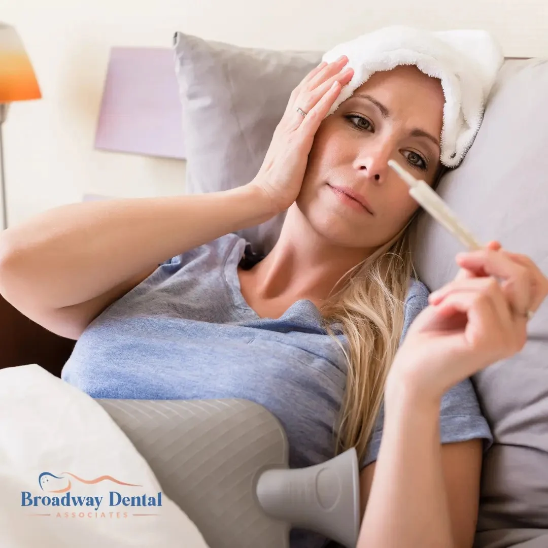 Woman in bed with a thermometer and a head compress, and Broadway Dental Associates logo in the bottom left of the image.