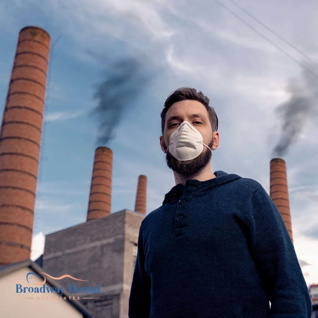 Man wearing a face mask by factory pollution and Broadway Dental Associates logo in the bottom left of the image.