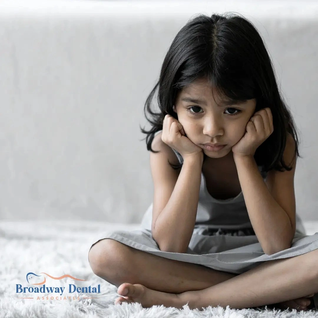 Sad young girl resting head on hands and Broadway Dental Associates logo in the bottom left of the image.