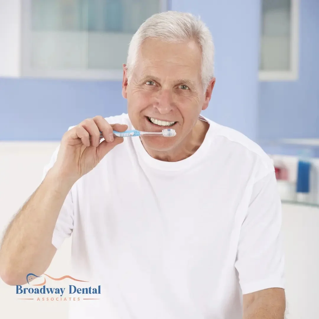 Senior man brushing teeth with smile and Broadway Dental Associates logo in the bottom left of the image.