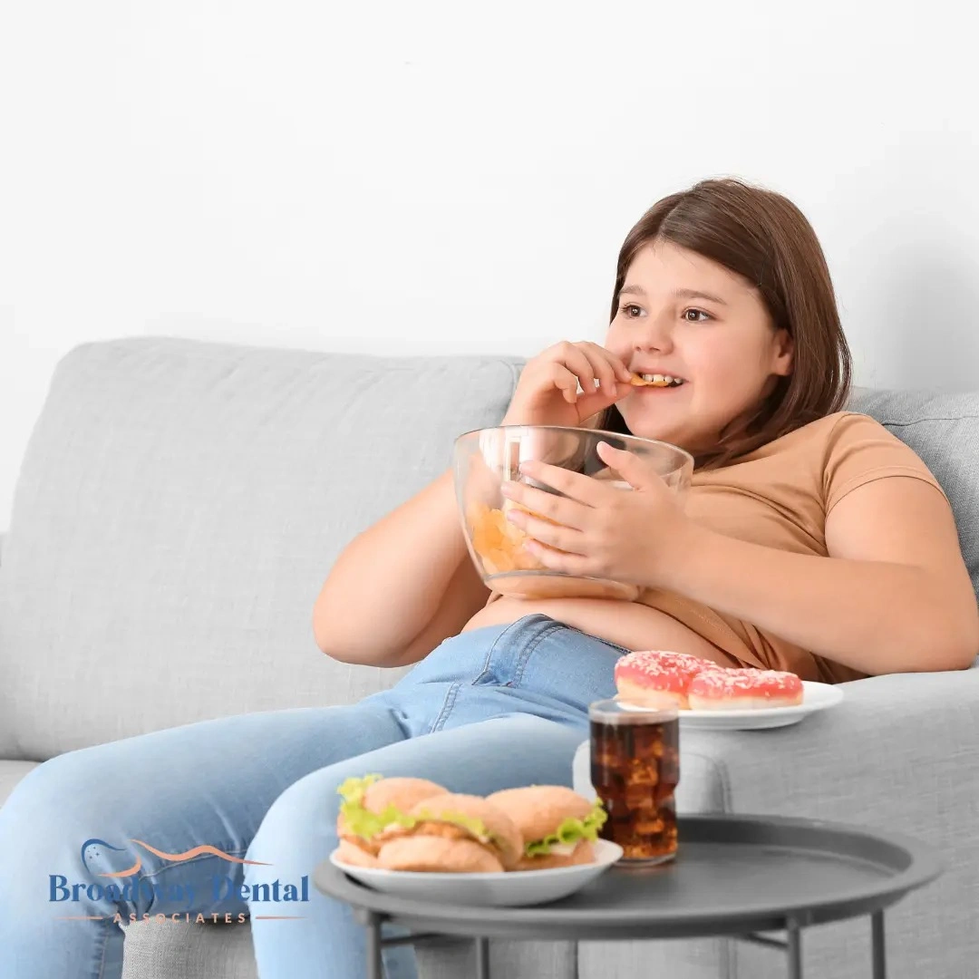 Young girl eating snacks on couch with junk food and Broadway Dental Associates logo in the bottom left of the image.